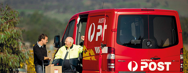 Two men talking in front of an Australia Post van. One man is leaning on a trolley with packages loaded on it.