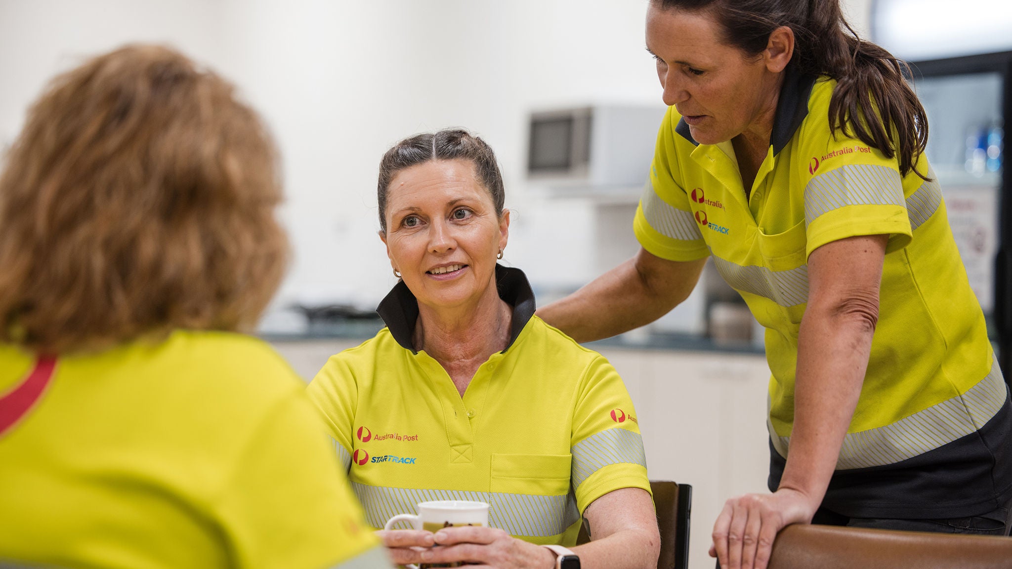 Three womon sitting around a table at an Australia Post facility wearing hi-vis uniforms