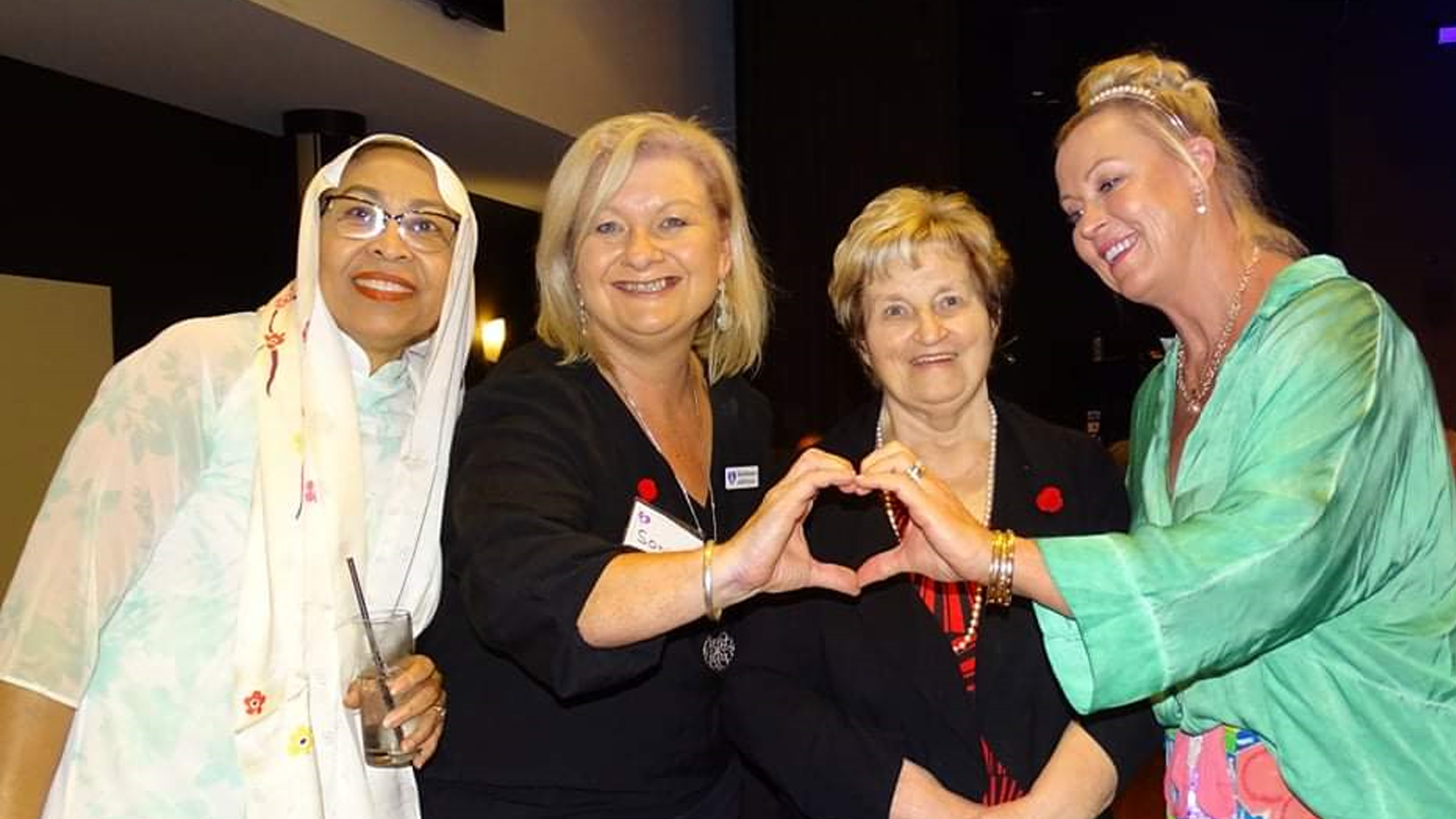 Four women are gathered together, with two of them forming a heart shape with their hands.