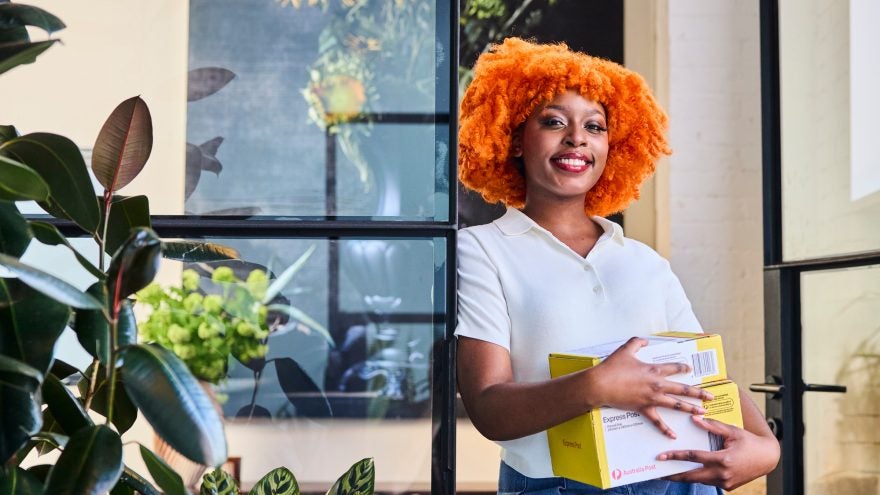 A woman with bright orange curly hair smiles while holding several yellow and white boxes. 
