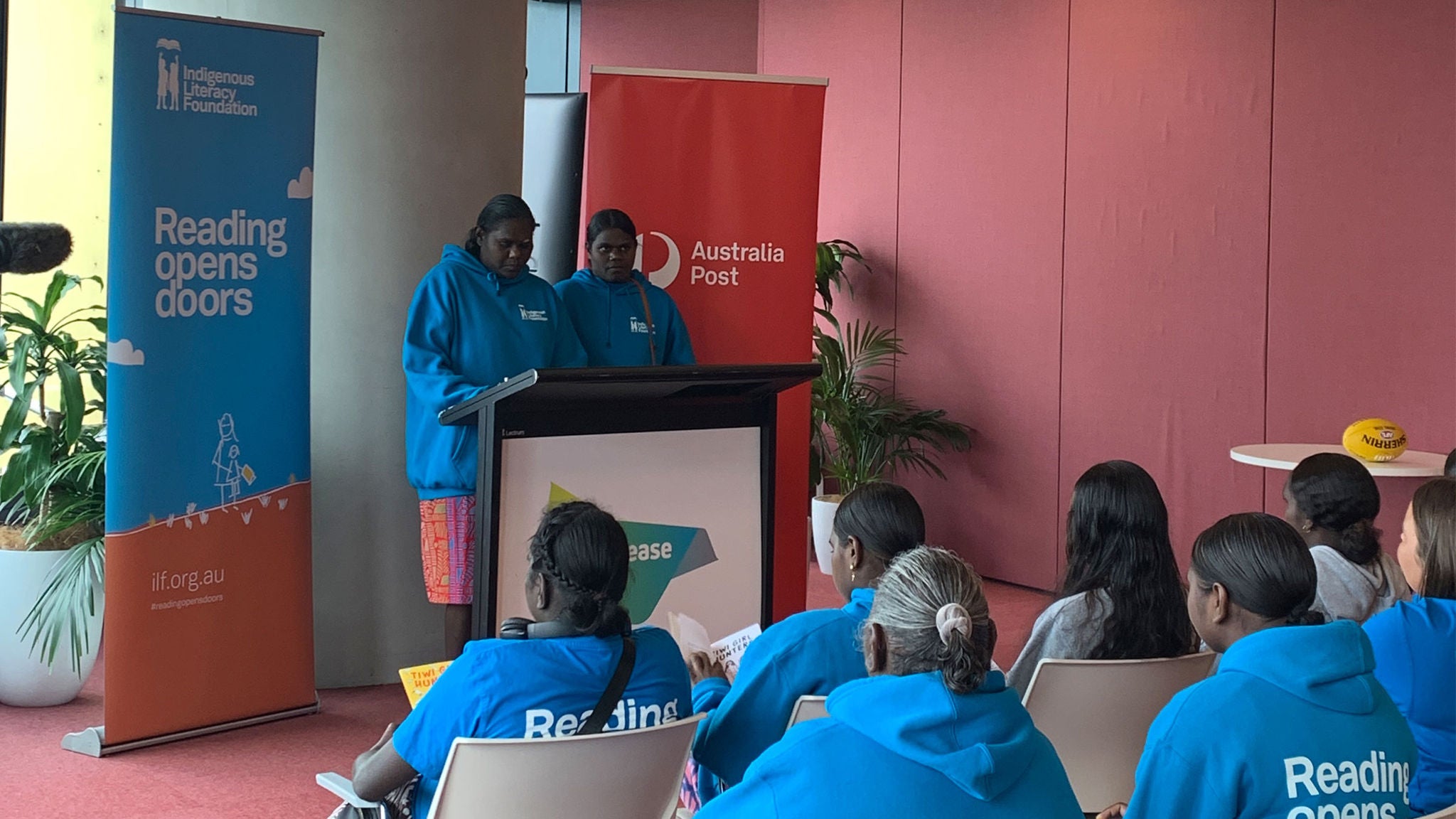 Two First Nations girls presenting in front of a group standing in front of indigenous literacy foundation and Australia Post banners