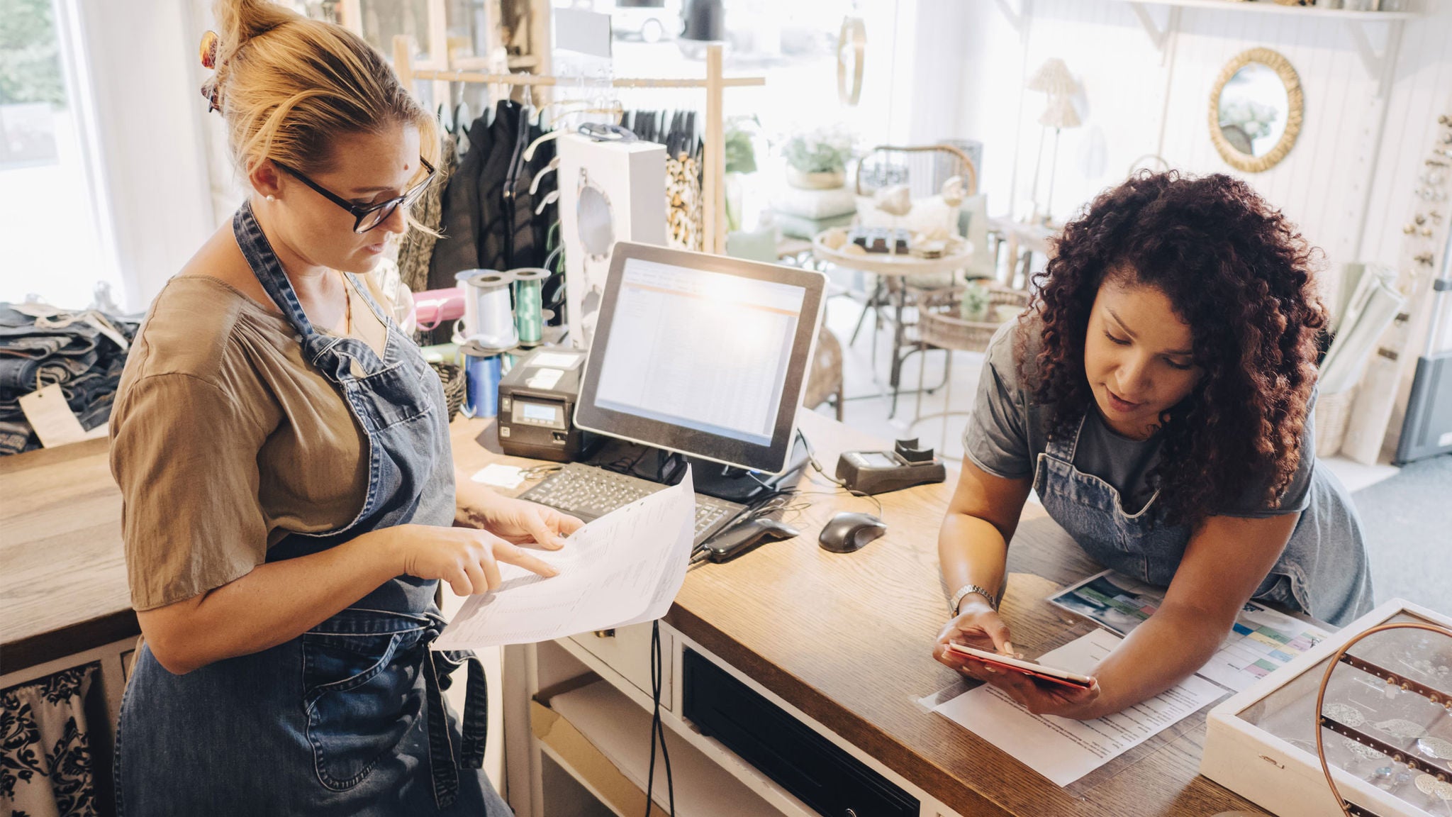 Two women are collaborating at a retail store counter, one reviewing paperwork while the other uses a smartphone.