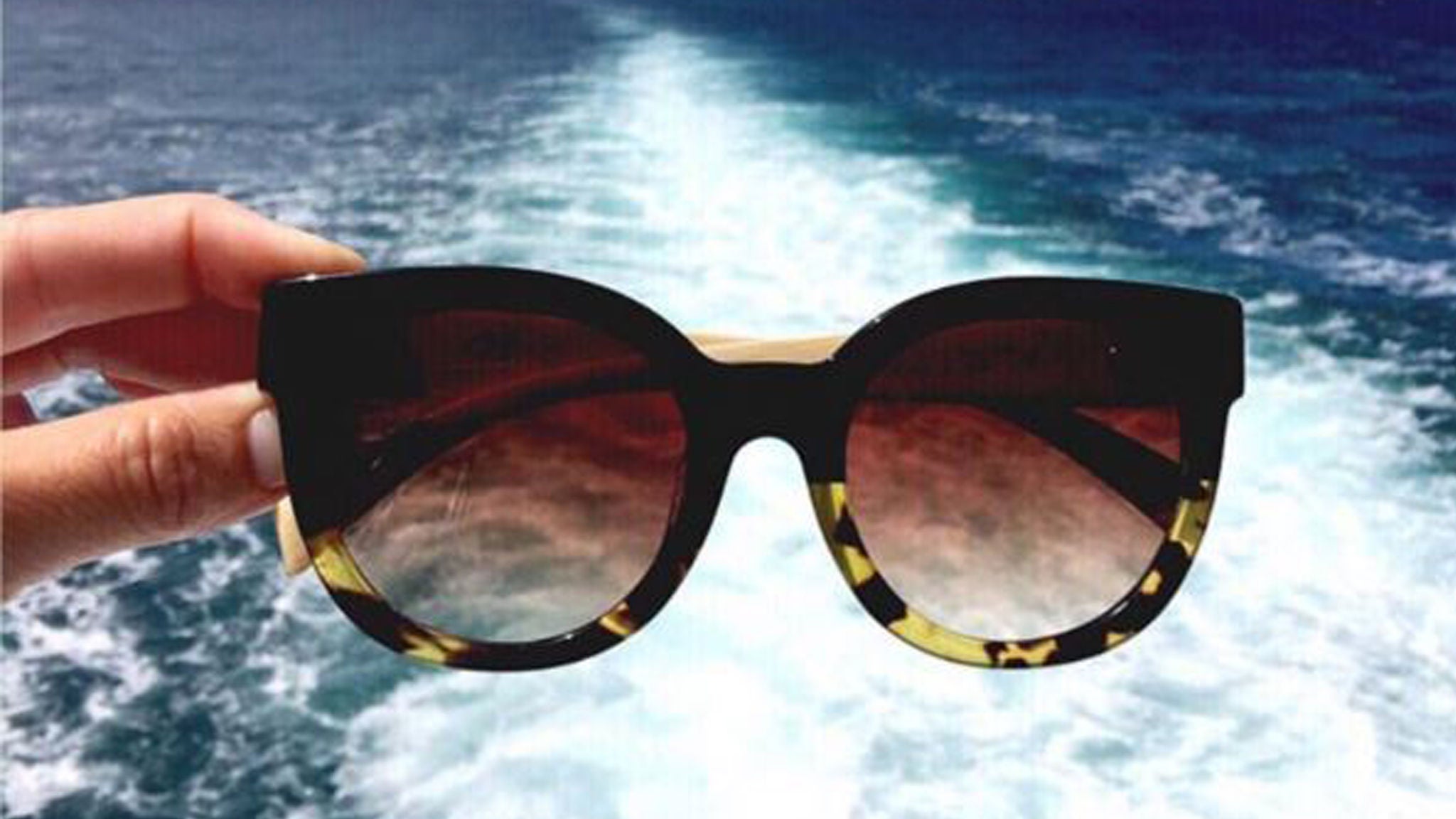 Hand holding Sticks and Sparrow sunglasses with ocean in background