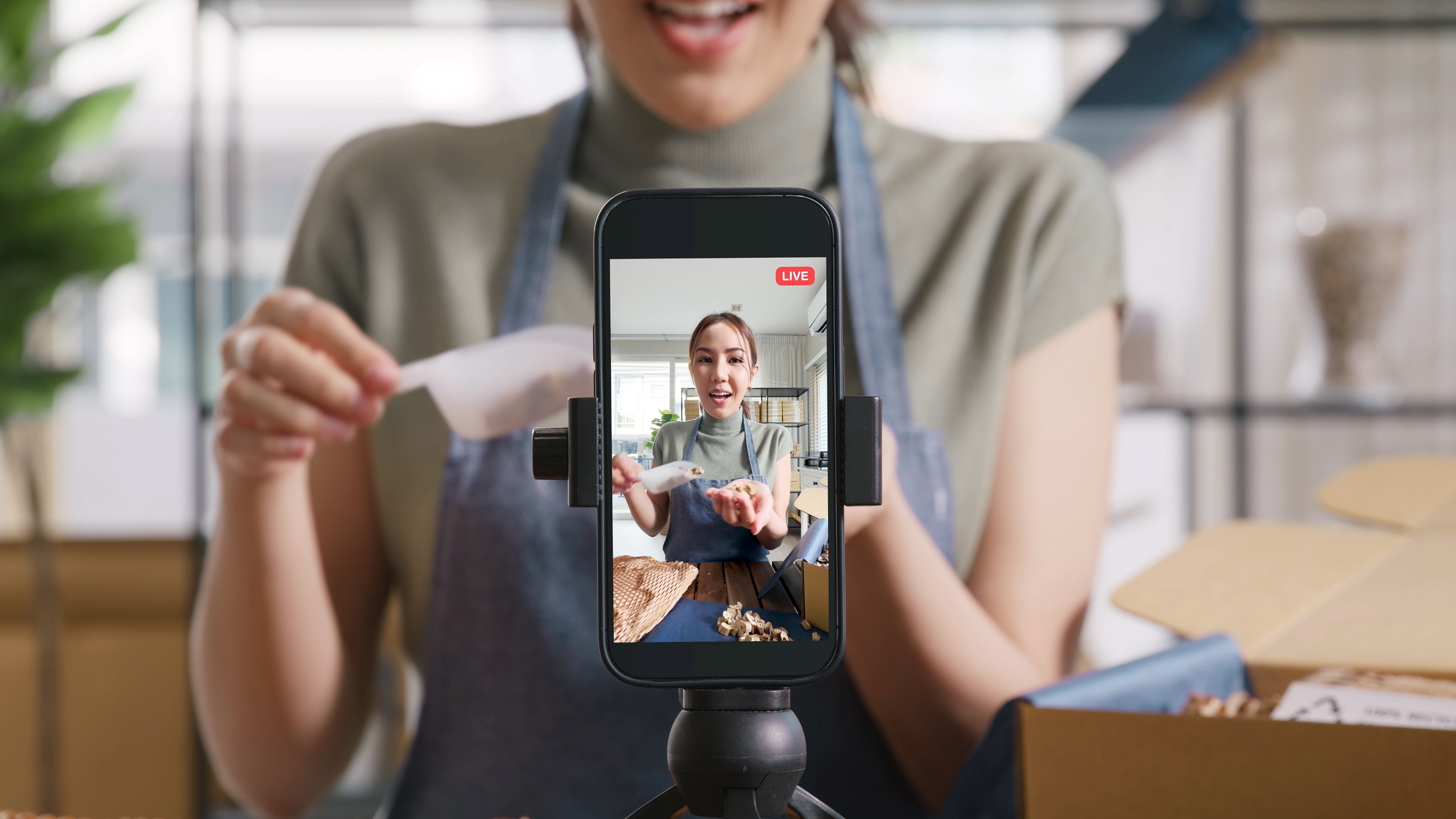 Smartphone filming a live shopping broadcast as a lady demonstrates a product during a livestream with packaging materials visible in the background.