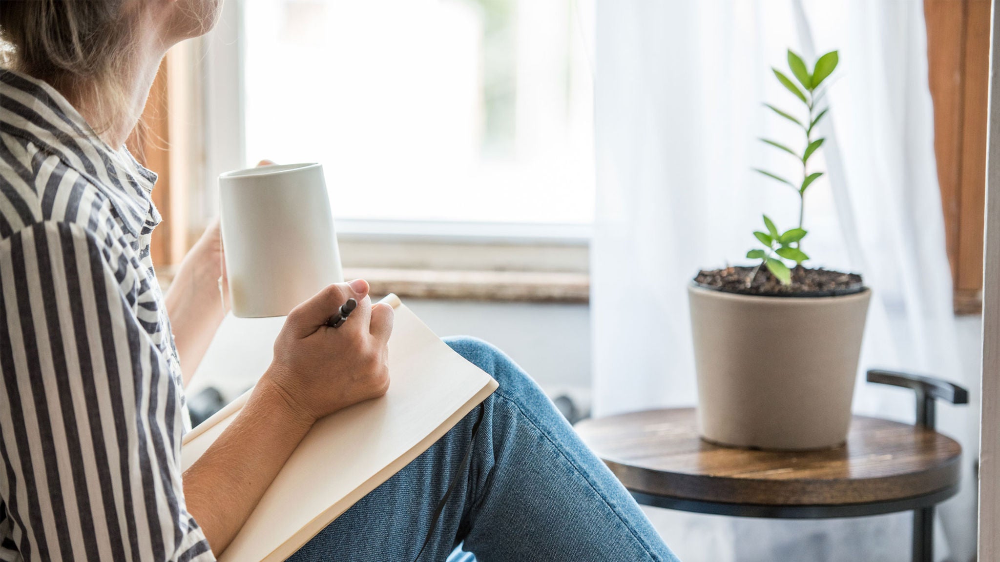 A woman sits indoors near a window, holding a white mug and writing in a notebook