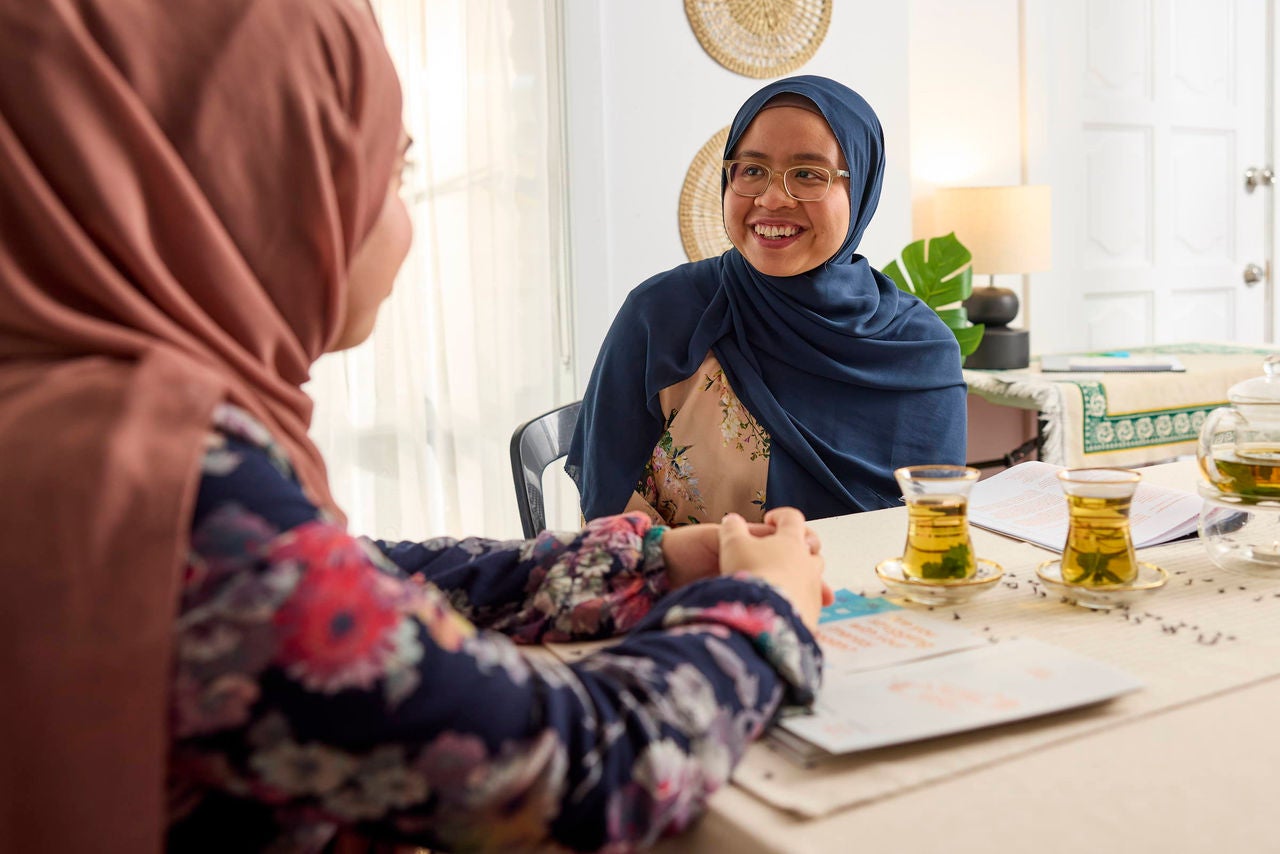 Two women wearing floral and solid-colored headscarves sit across from each other at a table in a room.