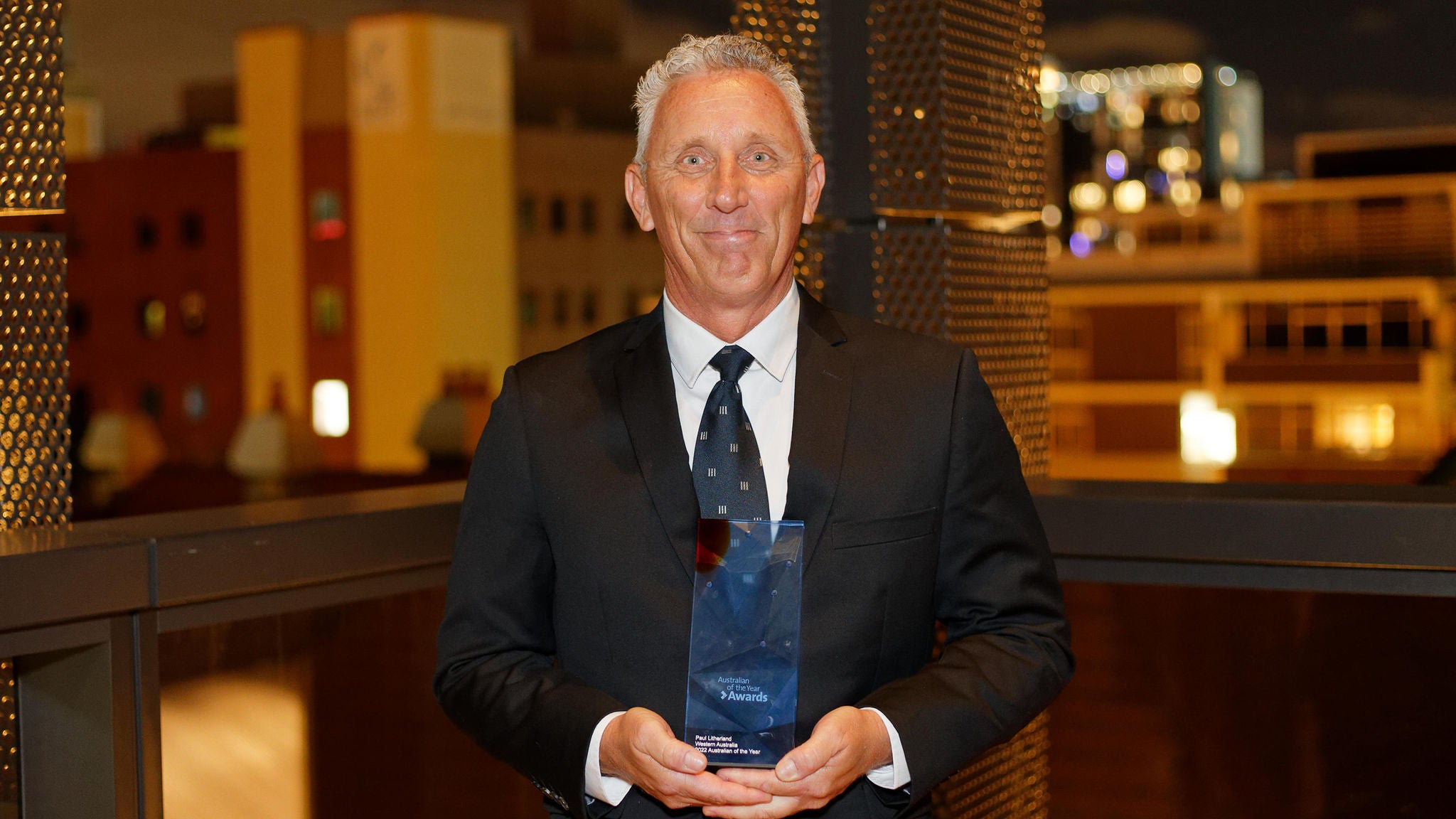 A smiling man in a dark suit holding the Australian of the Year Award 