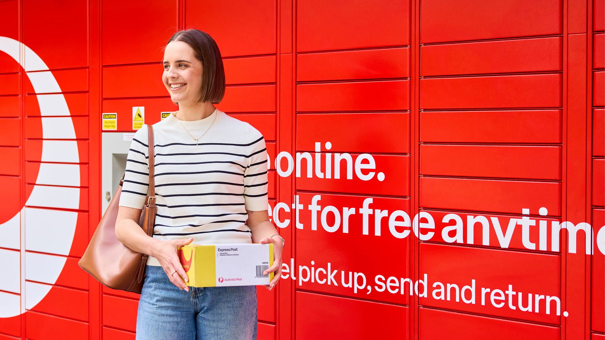 A woman stands in front of a red parcel lockers holding a small cardboard box