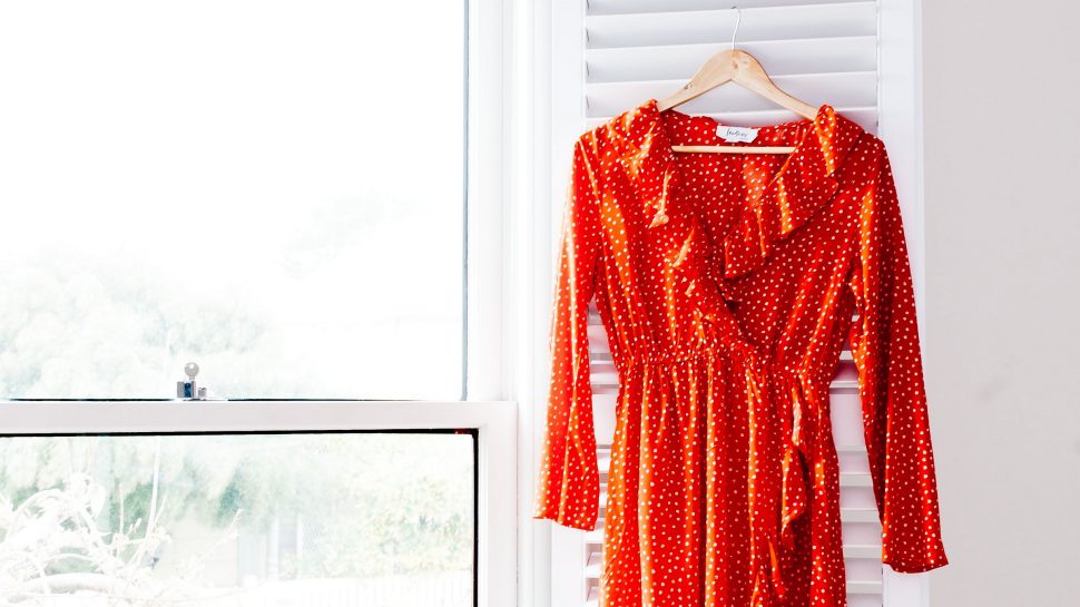 Woman standing by a window with a dress hanging up against a frame. The woman is holding the dress hem in one hand and smoothing the waist with her other hand - making sure the dress lays out nicely.