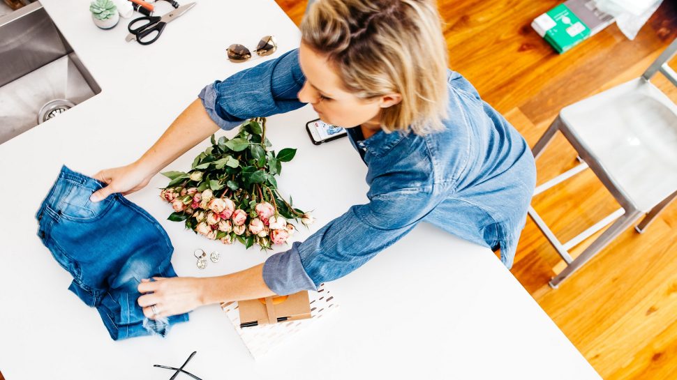 Woman leaning over a table with a bunch of flowers, sunglasses and a pair of jeans. She is adjusting the way the jeans are displayed, from neatly folded to a more relaxed look