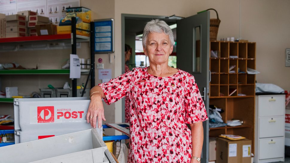 A white-haired woman in a white and red blouse standing with her right arm on an Australia Post plastic crate.  