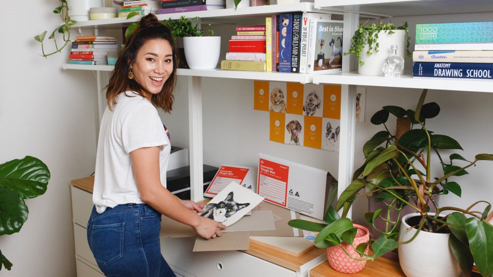 Jade Foo, founder of Creature & Cub, standing in her home office and holding a portrait of a dog. There are Australia Post envelopes on the table in front of her. 