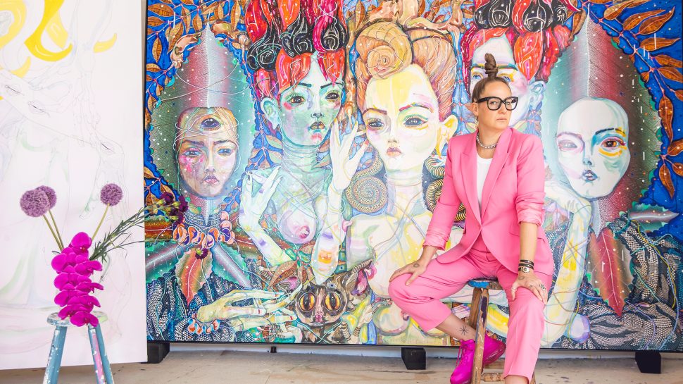  A woman wearing a pink suit and glasses sits on a stool and looks into the camera. Behind her is a large painted portrait of four women to her right is a vase of flowers on a wooden stool.