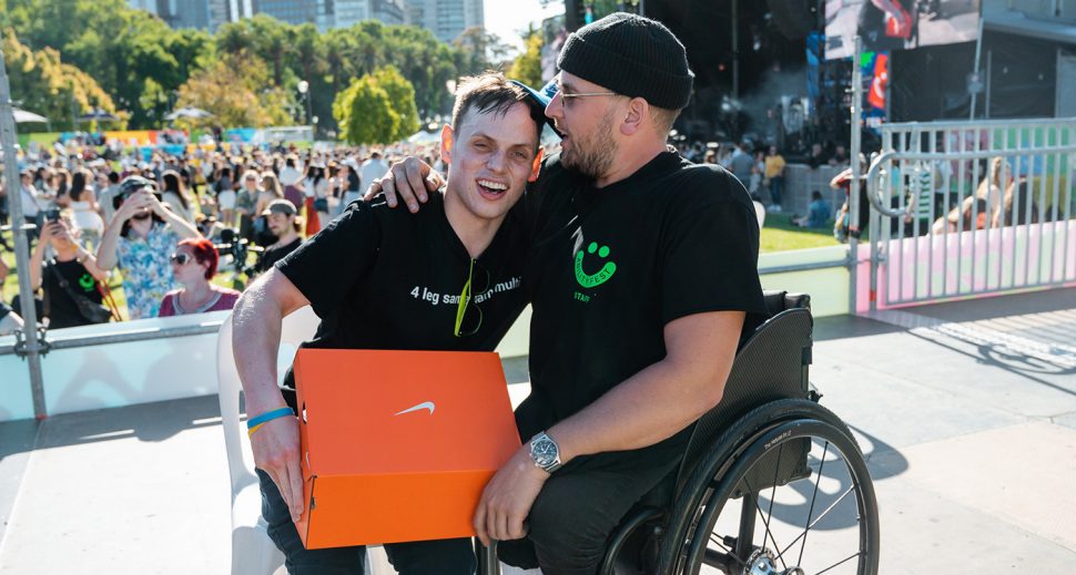 A man in a wheelchair has his right arm around a young smiling boy who is holding an orange Nike box. They are on stage with a crowd behind them. 