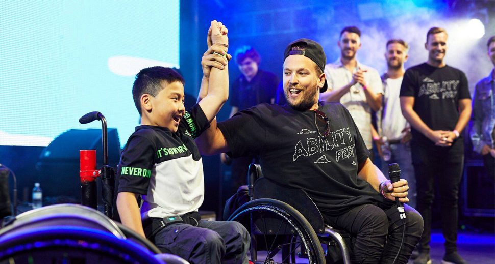 A man holding a young boy’s raised hands in celebration on stage. Both are in wheelchairs. 