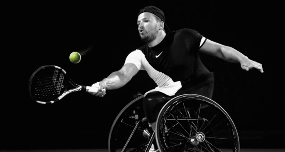 Black and white image of a man in a wheelchair hitting a tennis ball.
