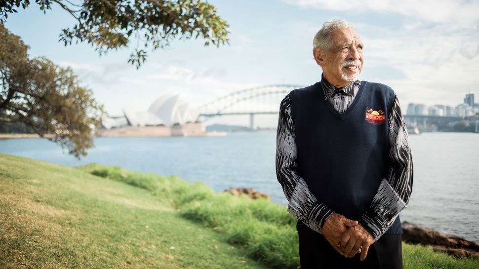 Gadigal Elder, Uncle Allen Madden, smiles at camera on Gadigal land with the Sydney Harbour Bridge in the distance behind him.