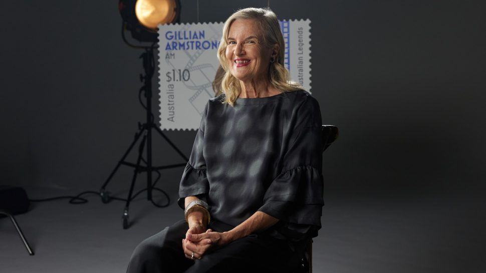 Gillian Armstrong sitting down at an interview, arms crossed, with the image of her Australia Post Legends of Filmmaking stamp in the background. 