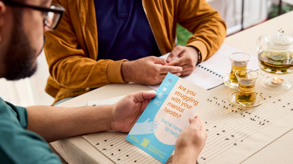 Over-the-shoulder view of a Muslim man holding a Helping Hands brochure. Text reads ‘Are you struggling with your mental health?’.