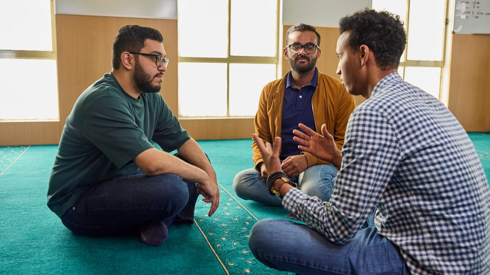Three young Muslim men sit cross-legged on the floor and enjoy a deep conversation. 