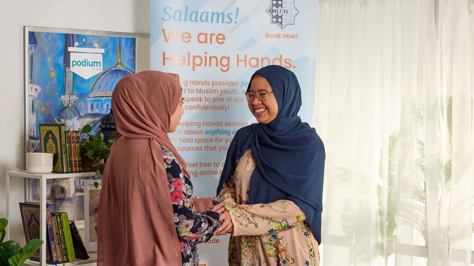Two Muslim women wearing hijabs smile and embrace in a handshake. Helping Hands signage is behind them. 