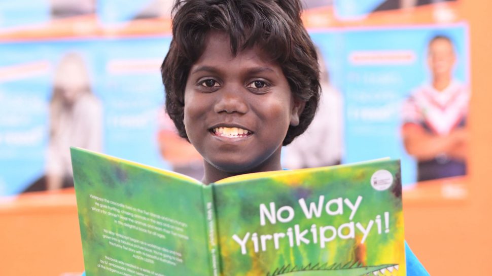 A smiling Aboriginal girl looks over a book titled ‘No Way Yirrikipay!