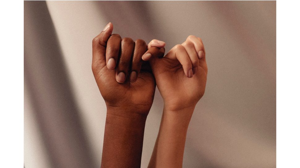 Two female hands, one with fair skin and one with dark skin, interlocking pinky fingers.