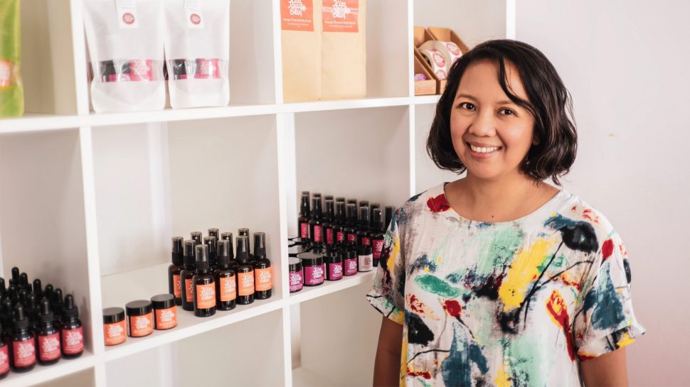 Hayley Worley, owner of The Sheet Society; Woman in white dress stands in front of a bed, smiling and holding a box.