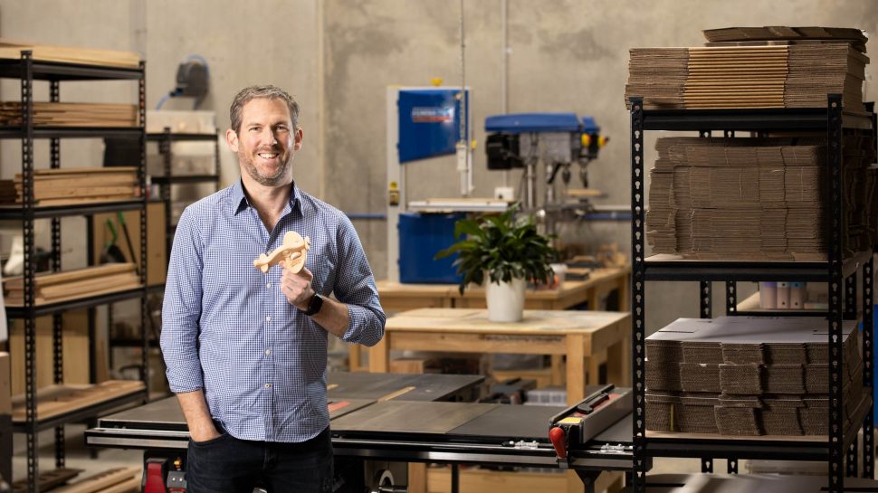 Man in a white shirt holding a wooden toy and standing against shelves of wooden slats. 