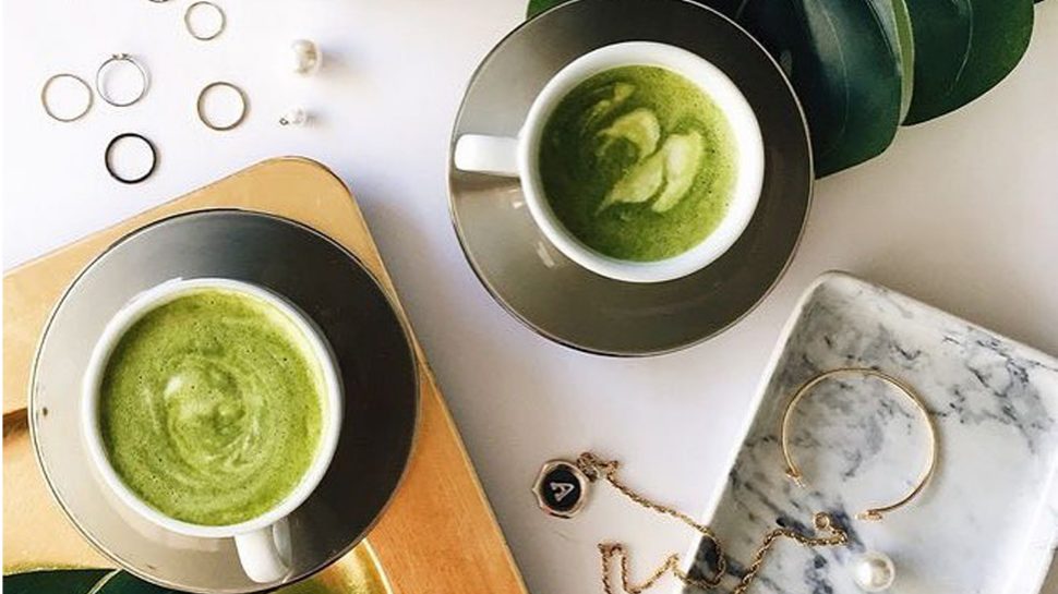 Two white coffee cups filled with matcha tea rest on a table with jewellery around it.