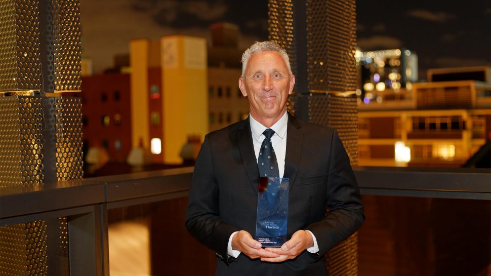 A smiling man in a dark suit holding the Australian of the Year Award 