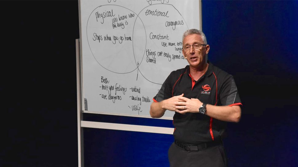A man in a black collared T-shirt delivering a presentation. There’s a whiteboard in the background. 