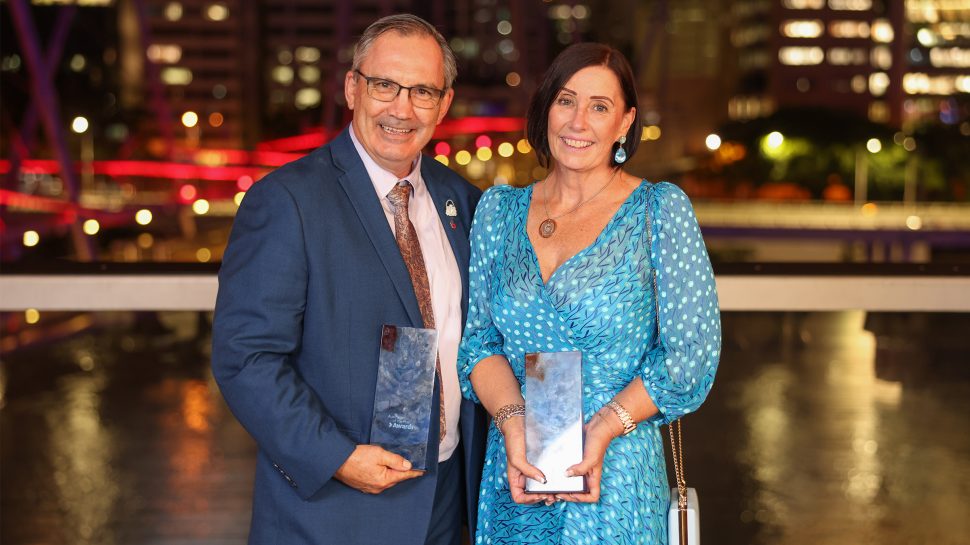 A man in a navy suit and a woman in a blue dress holding awards and smiling at the camera.