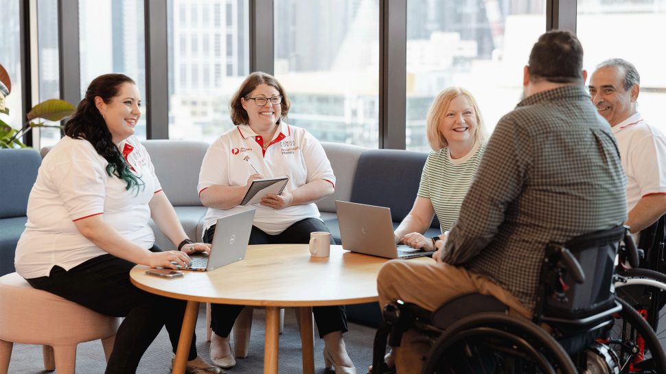 Three women and two men are sitting around a table talking. One of the men is in a wheelchair. 