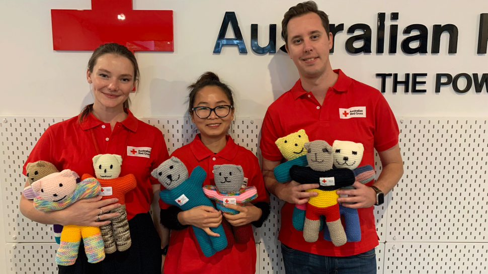 Two women and a man wearing Australian Red Cross uniforms and holding an armful of colourful knitted teddies.