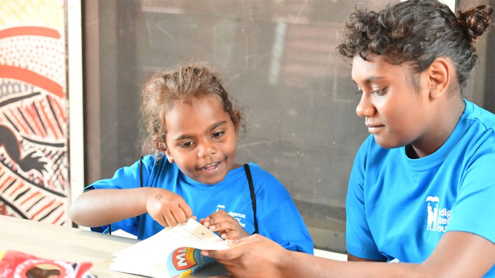 Two Aboriginal children wearing blue Indigenous Literacy Foundation t-shirts look at a book together.