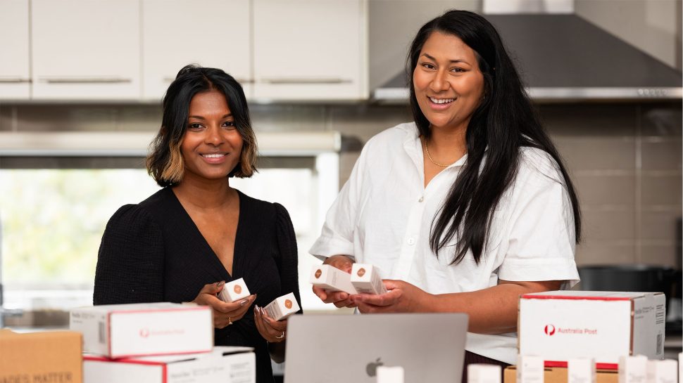 Two young olive-skinned women holding boxes of foundation and standing in front of a counter strewn with cosmetic products.