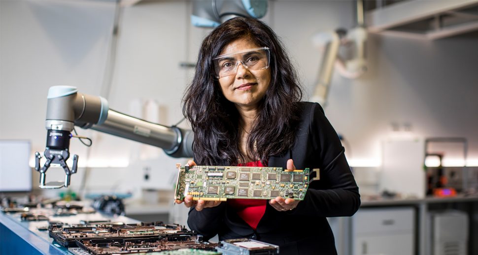 An olive-skinned woman in a red top, black blazer and protective eyewear standing in a lab and holding a circuit board. 