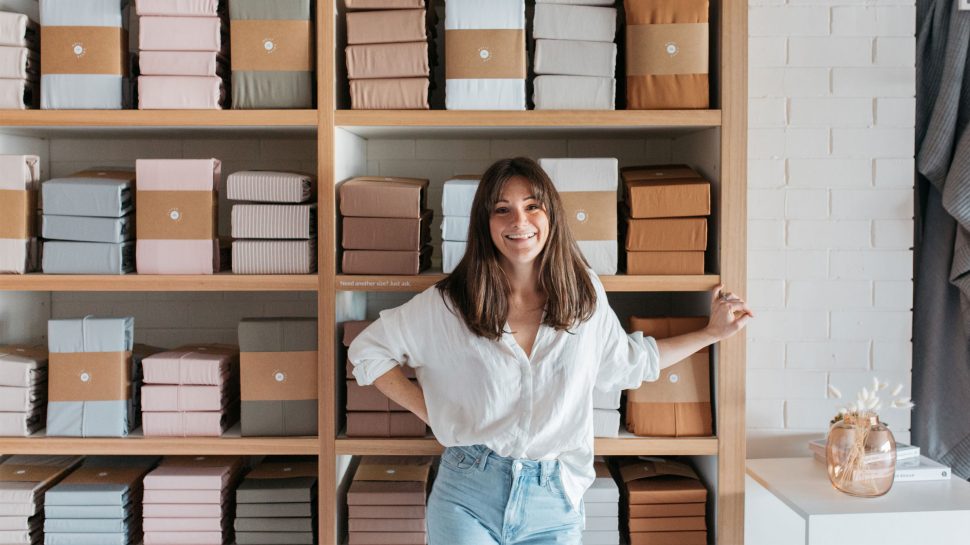 Hayley Worley, owner of The Sheet Society; A smiling young woman in a white shirt and blue jeans standing in front of tall wooden shelves stacked with various bed sheets. 