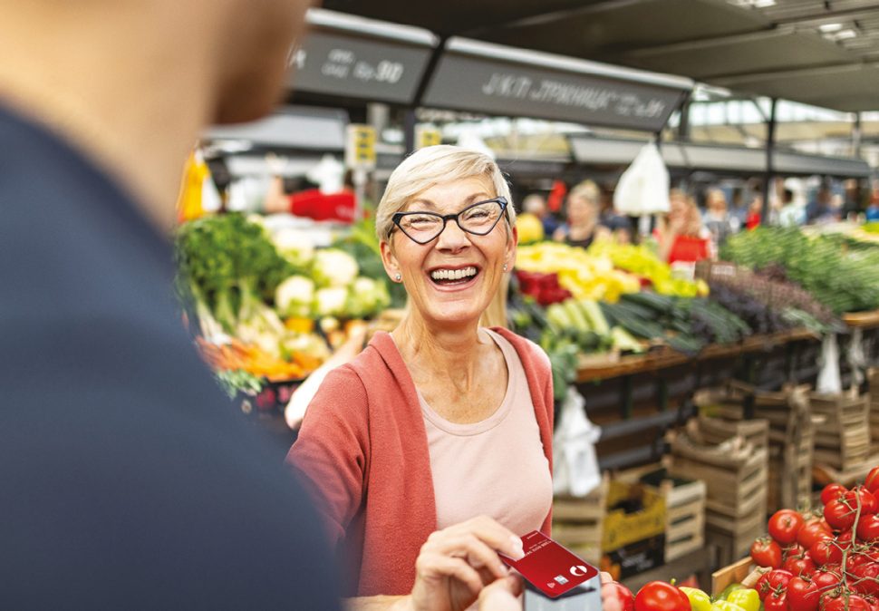 Woman tapping her Auspost Everyday Mastercard at the counter in a grocery store with the words "Budget, Shop and Save" written in the top left of the image.