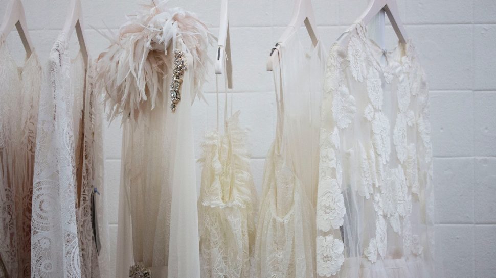 Six white dresses hang from a clothes rack in front of a white brick wall.