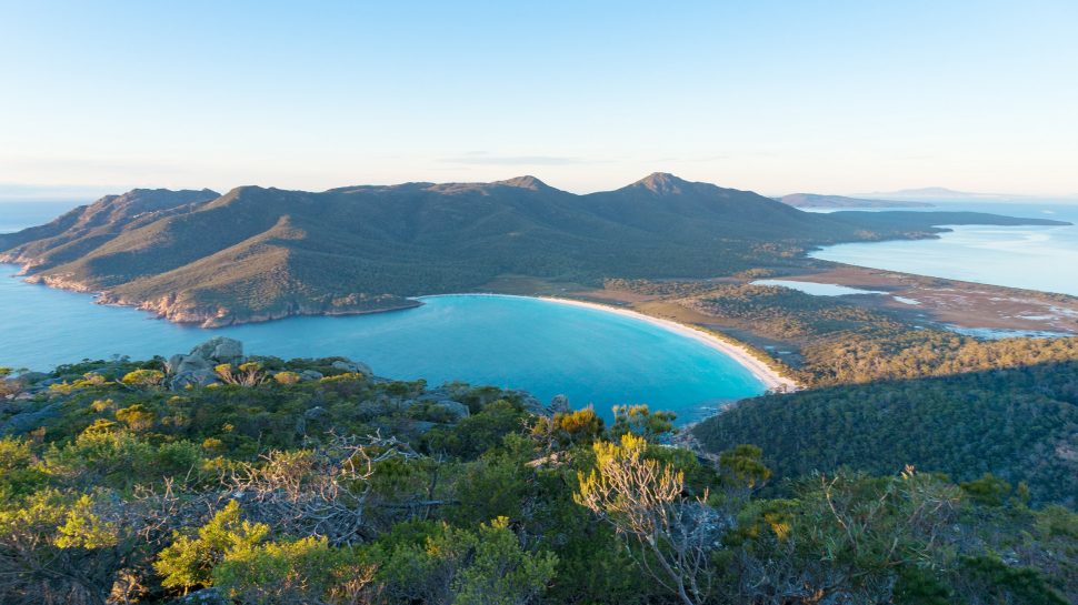 An aerial view of Wineglass Bay in Freycinet National Park, Tasmania. Calm water can be coming in from the ocean, forming the beautifully curved bay. On the right, the aqua water meets white sand and the sand joins a lush green forest. In the background there’s a cluster of green mountains with rocky cliff faces that meet the ocean.