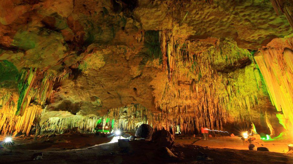 A panoramic view of Mammoth Cave in Kentucky. Stalactites hanging from a rocky ceiling makes up most of this scene. The stalactites hang low, and in the background they join with the ground. Green and yellow light from torches illuminate the cave. People can be seen on the right walking through the open space of the cave. Boulders - large and small - sit on the bottom of the cave floor. To the left you can see more torches shining brightly.