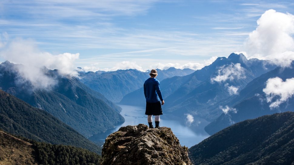 A man stands atop a peak in Fiordland National Park, looking out at the panoramic mountain view. 