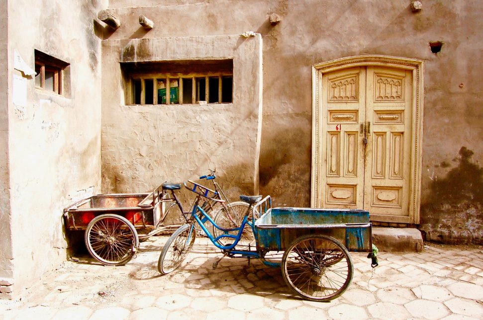 In a small sunlit cobblestone street in the town of Kashagr, two bicycles are parked outside the door to a building, with carts attached to the back. To the left, there is a corner wall with barred windows, and to the right, an arched doorway. 