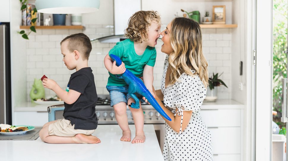Lady in a white summer dress with black polka dots laughing with a toddler wearing a green t-shirt and denim shorts and holding a blue toy dinosaur. He is standing on a kitchen counter with another toddler wearing a navy t-shirt and khaki shorts. 