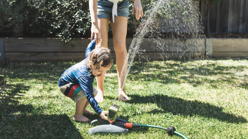 A drenched toddler wearing a blue long-sleeved top and colourful shorts plays with a garden sprinkler. A woman wearing a white shirt and denim shorts holds one of his hands