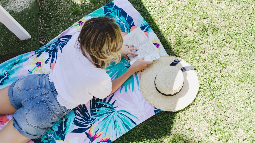 A lady is lying on a colourful towel reading a book. She’s wearing a white shirt and denim shorts, and has a summer hat beside her. 