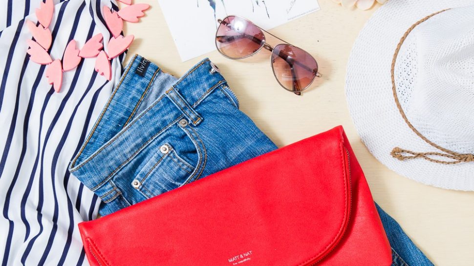 Flatlay photograph of a pair of jeans, a clutch, a shirt and necklace, sunglasses and a hat