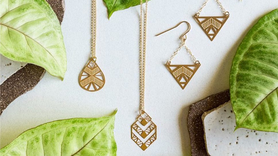 Two gold necklaces and a pair of earrings with an Aztec design draped in front of giant leaves.